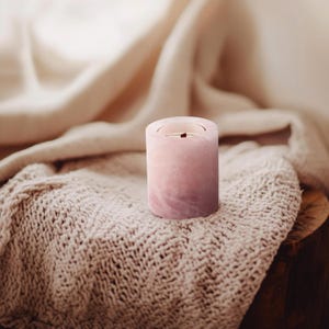 May include: A pink candle holder made of natural stone. The holder has a smooth, polished finish and a slightly textured surface. The candle holder is sitting on a soft, knitted blanket.