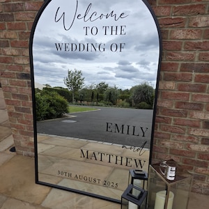 May include: A large, arched mirror with a black frame, displaying the text "Welcome to the Wedding of Emily and Matthew, 30th August 2025." The mirror reflects a cloudy sky and outdoor scenery. Two black lanterns and a silver lantern with candles are placed in front of the mirror.