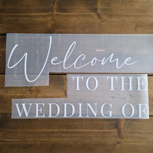 May include: White vinyl lettering spelling "Welcome TO THE WEDDING OF" on a clear transfer sheet. The lettering is in a mix of cursive and block fonts. The background is a dark brown wooden surface.