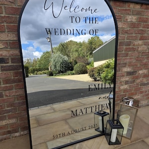 May include: A large, arched mirror with a black frame, reflecting a cloudy sky and a driveway. The mirror reads "Welcome to the Wedding of Emily and Matthew, 30th August 2025." Two black lanterns and a silver lantern with candles are placed in front.