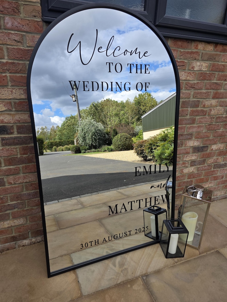 May include: A large, arched mirror with a black frame, reflecting a cloudy sky and a driveway. The mirror reads "Welcome to the Wedding of Emily and Matthew, 30th August 2025." Two black lanterns and a silver lantern with candles are placed in front.