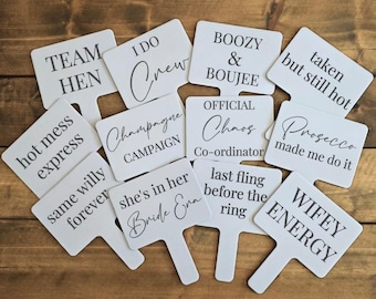 May include: A collection of white paddle signs with black text, arranged on a wooden surface. The signs feature phrases like "Team Hen," "I Do Crew," "Boozy & Boujee," and "Wifey Energy," ideal for a bachelorette party or bridal shower.