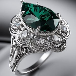 Puede incluir: Un anillo de plata con una piedra preciosa verde esmeralda en forma de pera rodeada de piedras preciosas más pequeñas y transparentes. El anillo tiene un diseño detallado y adornado.