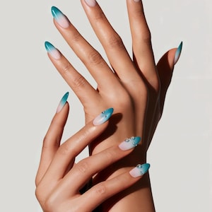 May include: Close-up of hands with long, almond-shaped nails painted with a gradient of teal and white. The nails are adorned with small, clear rhinestones.