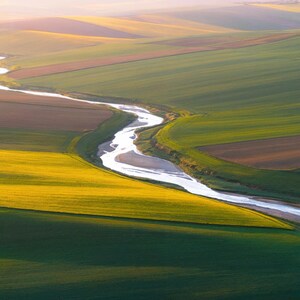 May include: Aerial view of a landscape featuring a winding river flowing through fields of varying colors. The fields are divided into sections of green, yellow, and brown, creating a patchwork pattern. The scene is bathed in soft sunlight.