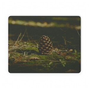 Pode incluir: Um pinha castanha repousa sobre uma superfície musgosa, rodeada por folhas caídas e galhos. A cena natural é capturada em close-up, com um fundo desfocado de árvores. A imagem é impressa em um mousepad retangular.