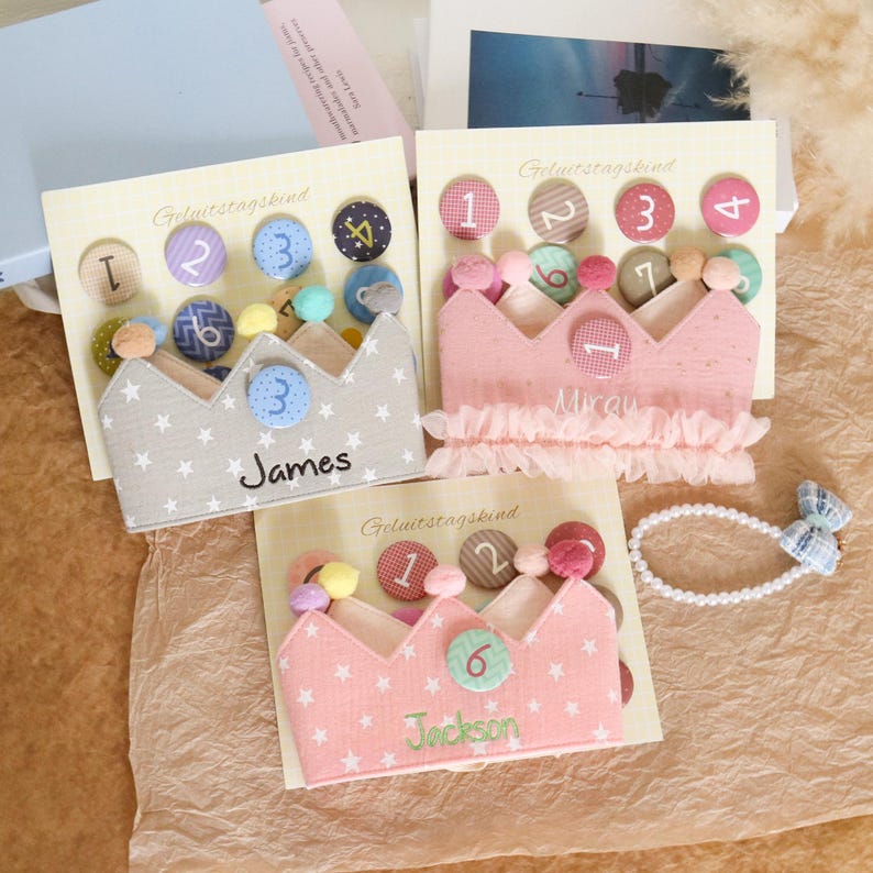May include: Three fabric crown headbands with star patterns and personalized names. Each crown has a number and decorative buttons. One is gray with white stars, one is pink, and the other is also pink.