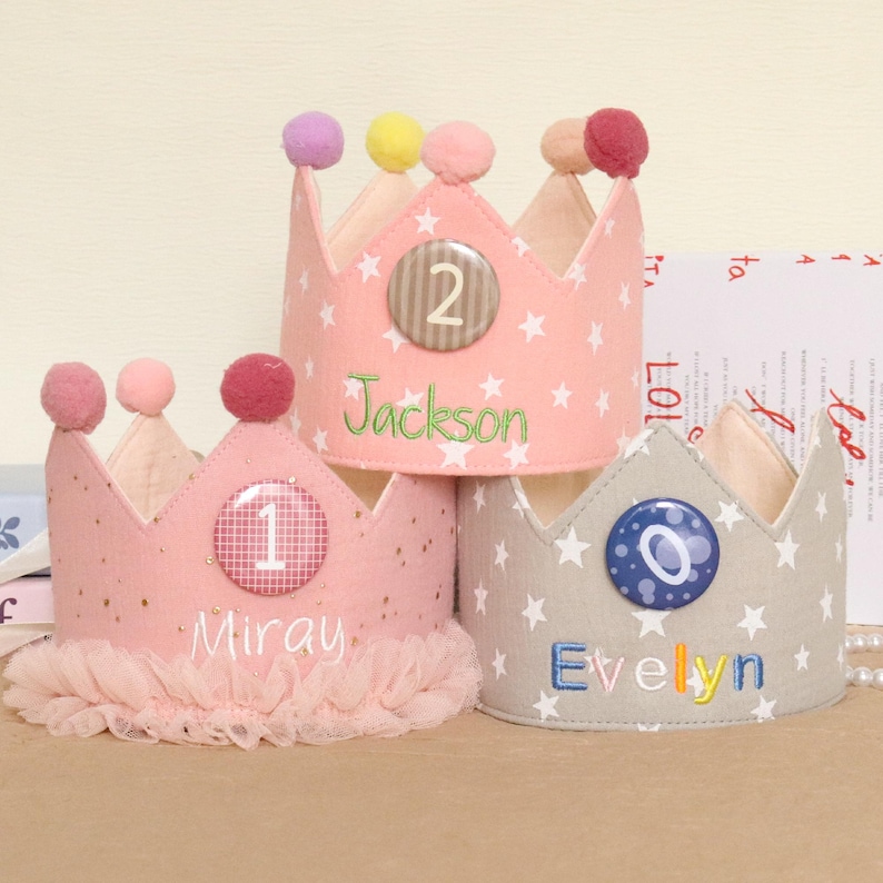 May include: Three handmade fabric crowns in pink and gray, decorated with stars, pom-poms, and personalized names and numbers. One crown has the name "Miray" and the number "1", another has "Jackson" and "2", and the third has "Evelyn" and "0".