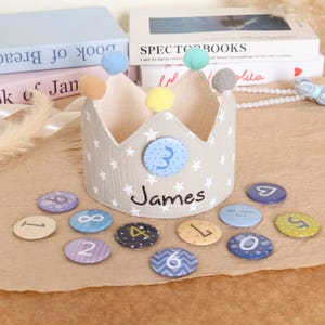 May include: A beige fabric crown with white stars, colorful pom-poms, and the name "James." Includes numbered circular badges. The crown has a blue badge with the number "3."