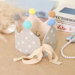 May include: A fabric crown with a star pattern and colorful pom-poms. The crown is light gray with white stars and has a cream-colored ribbon tie. The crown is decorated with gray, yellow, teal, and brown pom-poms. The crown is likely for a baby or child.
