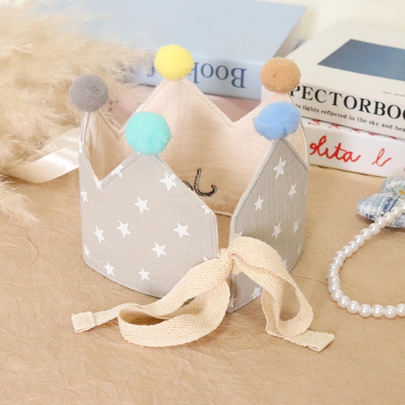 May include: A fabric crown with a star pattern and colorful pom-poms. The crown is light gray with white stars and has a cream-colored ribbon tie. The crown is decorated with gray, yellow, teal, and brown pom-poms. The crown is likely for a baby or child.