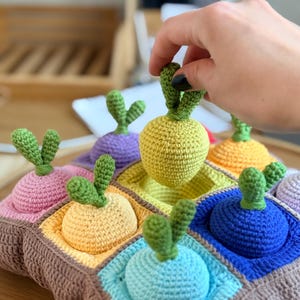 May include: A colourful crochet toy featuring a brown base with multiple square pockets, each holding a crocheted radish. The radishes are in various colours, including yellow, pink, purple, and blue, with green leafy tops. A hand is holding one of the radishes.