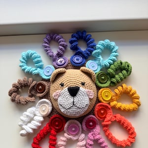 Può includere: Una testa di orso all'uncinetto con viso color marrone chiaro, muso bianco e naso nero, circondata da dodici elastici per capelli all'uncinetto a spirale di vari colori, ognuno con un bottone in plastica abbinato al centro dell'anello.