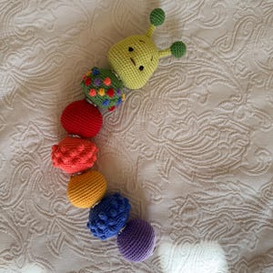 May include: A multi-coloured crochet caterpillar toy featuring a light green head with two antennae, followed by a series of connected spherical segments in green with colourful dots, red, textured orange, yellow, textured blue, and purple.