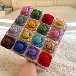 May include: A square crochet sensory mat featuring a grid of sixteen colorful squares, each with a raised circular center. The squares are joined by white yarn and include shades of red, blue, green, yellow, brown, purple, gray, and pink.