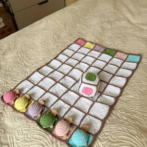 Peut inclure: Une couverture pour bébé au crochet faite à la main avec un motif quadrillé blanc et des bordures marron. La couverture est ornée de tortues au crochet colorées le long d'un bord et d'un jouet cube assorti.