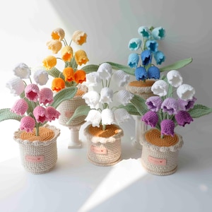 Pode incluir: Uma coleção de flores de lírio-do-vale feitas à mão em crochê em várias cores, incluindo branco, rosa, amarelo, laranja, azul e roxo. Cada flor está em um vaso de crochê bege com folhas verdes.
