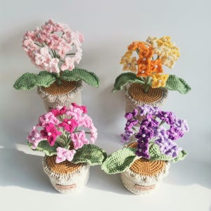 Puede incluir: Cuatro arreglos florales de ganchillo en macetas. Las flores son en tonos de rosa, naranja, amarillo y morado, con hojas verdes. Cada flor tiene detalles de perlas. Las macetas son beige y marrón.