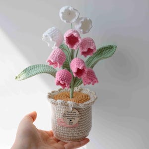 May include: A handmade crochet flower arrangement in a beige pot. The arrangement features pink and white bell-shaped flowers, green leaves, and a beige pot with a scalloped edge. A small tag is attached to the pot.