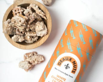 May include: A cylindrical orange container with a blue and white label that reads "GEAUX NUTS New Orleans Style Treats WHITE CHOCOLATE CRUNCH Meringue pecans, crunchy grahams, and pretzels roasted in white chocolate Net wt 6 oz / 170g". The container is decorated with a pattern of blue trumpets on an orange background. A bowl of white chocolate covered pretzels, pecans, and graham crackers is in the background.