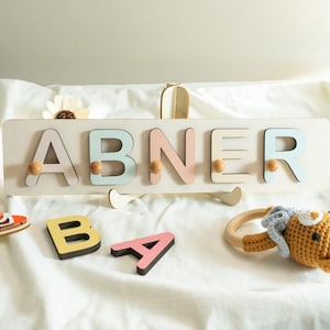 Puede incluir: Un rompecabezas de nombre de madera con el nombre "ABNER" en colores pastel. Las letras están unidas a una tabla de madera color crema con clavijas. Letras de madera adicionales "B" y "A" están cerca, junto con un juguete de madera y una sonaja de ganchillo.