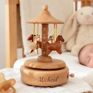 May include: A wooden carousel music box with four horse figurines. The carousel has a light brown finish and the name "Micheal" is engraved on the base. The carousel is set on a white surface, with a pacifier in the foreground.