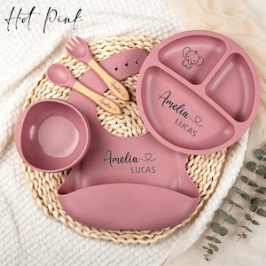 May include: A hot pink silicone feeding set including a three-sectioned plate, a bowl, a bib, and a wooden-handled spoon and fork. The plate and bib are personalised with the name Amelia Lucas and a heart icon. The plate features an elephant graphic.