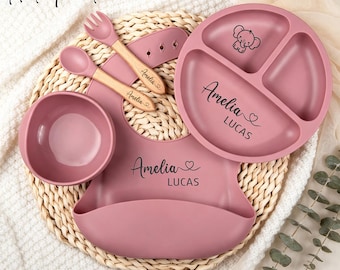 May include: A hot pink silicone feeding set including a three-sectioned plate, a bowl, a bib, and a wooden-handled spoon and fork. The plate and bib are personalized with the name Amelia Lucas and a heart icon. The plate features an elephant graphic.