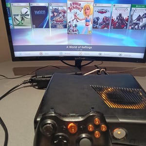 May include: A black Xbox 360 console and controller. The screen shows a game menu, including "Assassin's Creed Brotherhood" and "A World of Keflings". The controller's buttons are lit up. The console is on a surface.