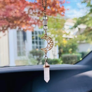 May include: A hanging ornament featuring a gold-toned metal crescent moon charm with star cutouts, suspended from a chain with three clear and pale pink beads. A faceted, light pink crystal point hangs from the bottom of the crescent moon charm.