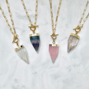 Puede incluir: Cuatro collares con cadenas de eslabones rectangulares dorados y cierres de barra. Cada collar tiene un colgante de piedra tallada en punta con una tapa de metal dorado. Los colgantes son transparentes, morados y verdes, rosa claro y blanco translúcido.