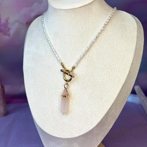 May include: A delicate necklace featuring a strand of small white pearls. The necklace has a gold-colored toggle clasp and a pendant with a pink crystal. The crystal is encased in a gold-colored setting. The necklace is displayed on a white mannequin.
