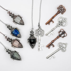 May include: A collection of four teardrop-shaped gemstone pendants on silver chains, featuring white, blue, pink, and green stones in ornate metal settings, alongside a silver key pendant and four decorative metal keys in copper and silver tones.
