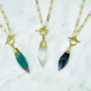 May include: Three gold-toned necklaces with gemstone pendants. Each necklace features a toggle clasp and a chain. The pendants are in shades of teal, clear, and purple, with gold-toned caps. The necklaces are displayed on a white marble surface.