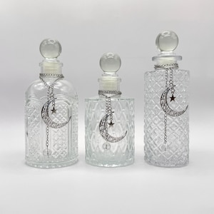 May include: Three clear glass bottles with spherical stoppers and textured diamond patterns. Each bottle features a silver-toned chain necklace adorned with a filigree crescent moon charm and a small star, hanging from the neck of the glass vessel.