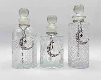 May include: Three clear glass bottles with spherical stoppers and textured diamond patterns. Each bottle features a silver-toned chain necklace adorned with a filigree crescent moon charm and a small star, hanging from the neck of the glass vessel.