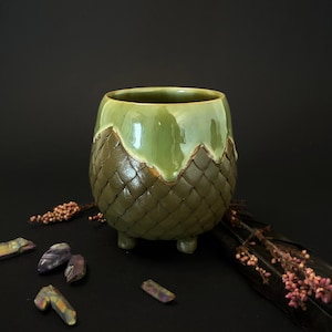 May include: A ceramic mug with a rounded body and three small feet. The upper portion features a smooth, glossy light green glaze, while the lower section has a textured, dark olive green scale pattern. The mug is surrounded by small crystals.