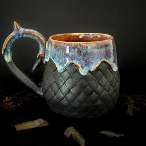 Könnte beinhalten: Handgefertigte Keramiktasse mit einem einzigartigen Drachenschuppen-Design. Die Tasse hat einen schwarzen, strukturierten Boden, einen braunen Innenraum und einen blau-violett glasierten Rand. Der Griff ist flossenartig geformt.