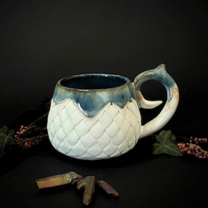 May include: A handmade ceramic mug with a white, textured body and a blue rim and handle. The mug has a unique scale-like pattern and a curved handle. The interior is a deep blue. Decorative crystals and foliage are in the background.