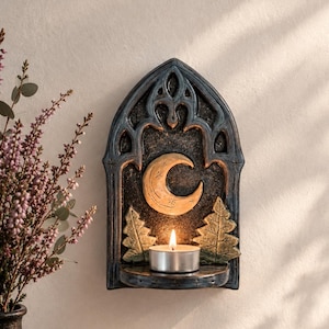 Pode incluir: Um suporte de vela de parede em estilo gótico escuro com uma moldura em arco pontiagudo, um relevo central de lua crescente e duas árvores perenes estilizadas. Uma pequena vela de chá acesa repousa na prateleira integrada abaixo da lua.