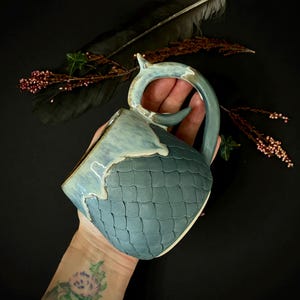 May include: A handmade ceramic mug with a textured, teal-colored exterior and a light blue rim and handle. The mug has a unique, scale-like pattern on its body and a distinctive handle design. The mug is held against a dark background, with decorative elements.