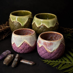 Könnte beinhalten: Vier Keramikbecher mit einem strukturierten Schuppenmuster auf der Außenseite. Zwei Becher haben eine grüne Glasur mit einem helleren grünen Rand, während die anderen beiden eine violette Basis mit einer cremefarbenen Glasur oben haben.