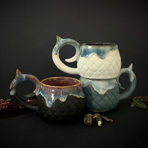 Könnte beinhalten: Drei Keramikbecher mit einem Fischschuppen-Design. Die Becher sind in Blau-, Weiß- und Schwarztönen gehalten und haben einzigartige Griffdesigns. Die Becher sind übereinander gestapelt. Die Becher stehen auf einer schwarzen Oberfläche.