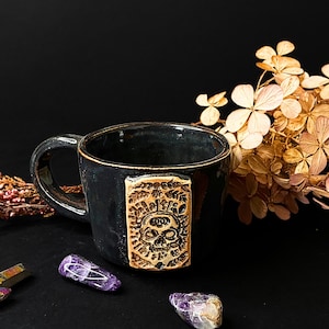Può includere: Una tazza in ceramica nera con una placca rettangolare in rilievo color beige sulla parte anteriore. La placca presenta un design in rilievo di un teschio circondato da intricati motivi a foglia. La tazza ha una finitura lucida.