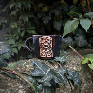 Könnte beinhalten: Dunkelblaue Keramiktasse mit Henkel und einem geschnitzten Totenkopf-Design auf einem rechteckigen Feld. Die Tasse steht vor grünem Laub auf einer Steinoberfläche.