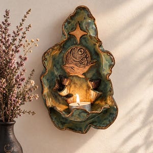 Pode incluir: Um castiçal de parede em cerâmica com esmalte manchado em tons de azul-petróleo e castanho. A peça apresenta um relevo de uma baleia e uma estrela de quatro pontas. Uma pequena prateleira inferior sustenta uma vela acesa e detalhes em conchas.