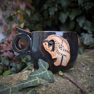 Puede incluir: Taza de cerámica negra hecha a mano con un diseño de asa único. La taza presenta un relieve tallado, de color marrón claro, de un animal estilizado. La taza está sobre una superficie de piedra con hojas verdes en primer plano.