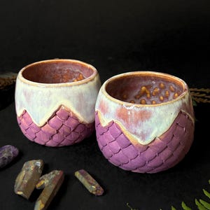 Puede incluir: Dos tazas de cerámica hechas a mano con un diseño de escamas de dragón. Las tazas son moradas con un borde blanco y crema. El interior de las tazas es de color marrón rojizo. Cristales y un helecho en el fondo.