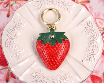 Puede incluir: Un llavero de cuero rojo en forma de fresa con una hoja de cuero verde y un anillo de llave de metal dorado.