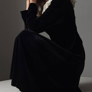 Pu&ograve; includere: Abito lungo nero in velluto con colletto in pizzo bianco. L'abito ha maniche lunghe e una gonna fluida. La modella &egrave; seduta, con le mani vicino al viso. L'abito &egrave; uno stile classico ed elegante.